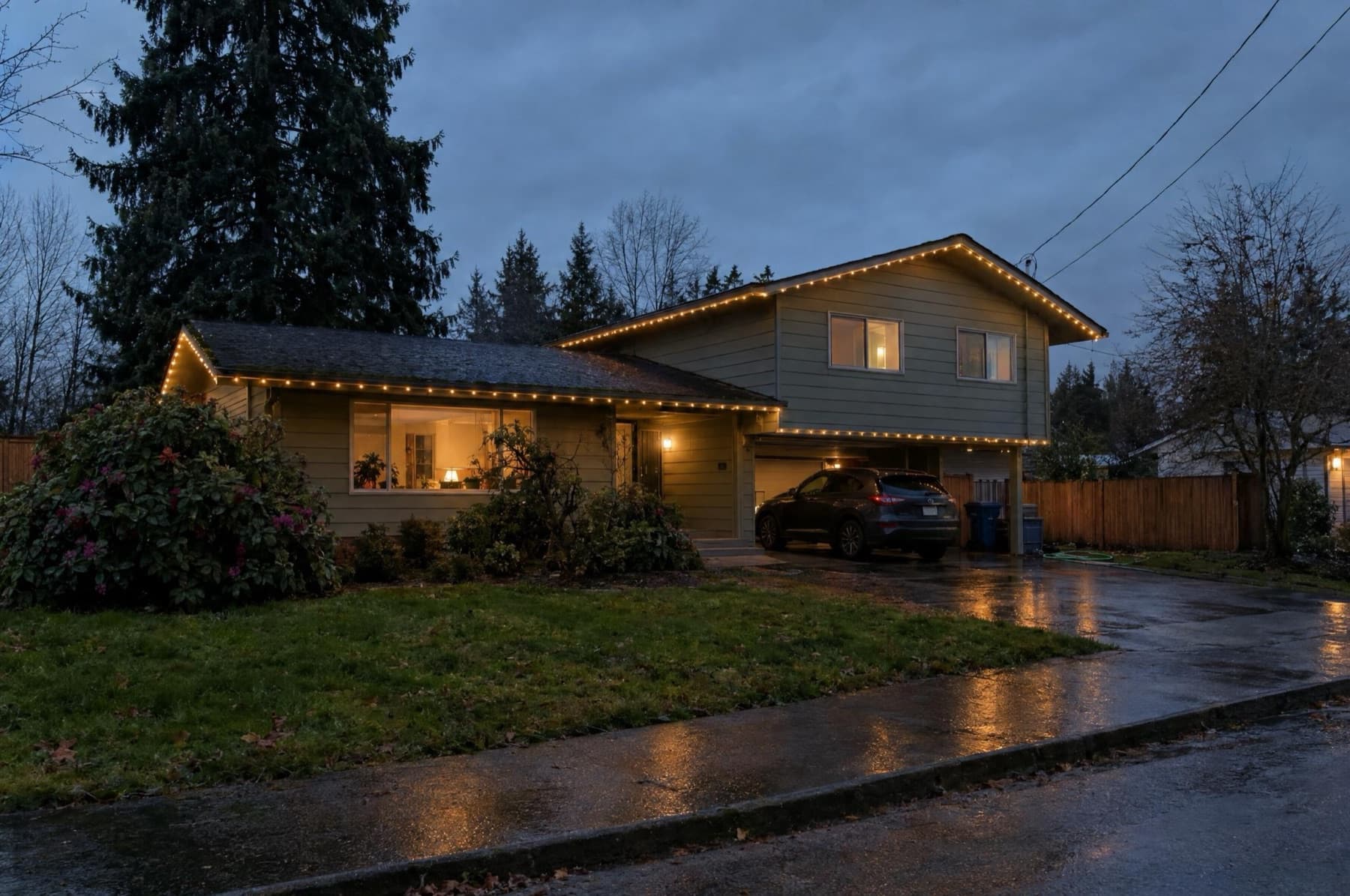 1960s split-level with both rooflines lit warm white, rhododendrons in front