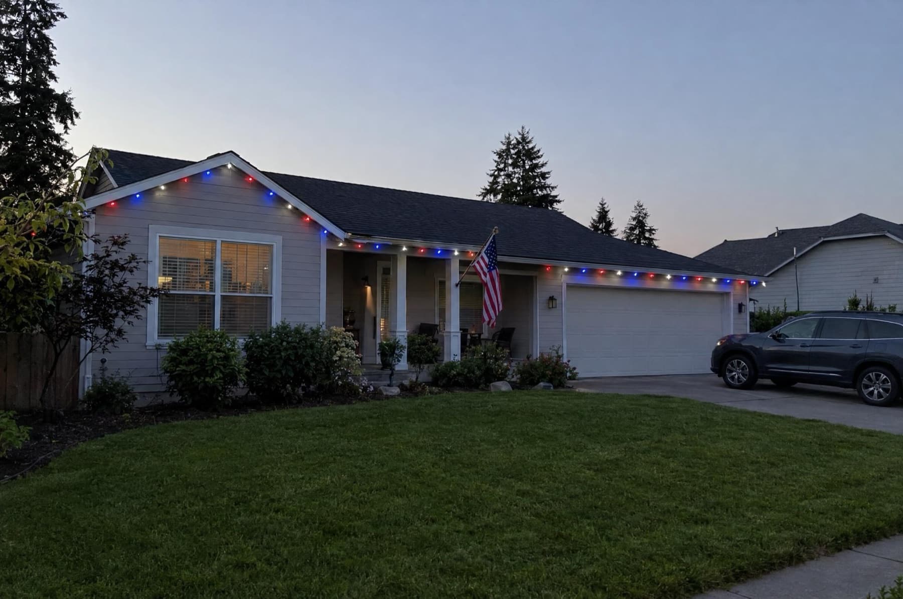 Ranch house at July dusk with red, white and blue roofline lights and a flag on the porch