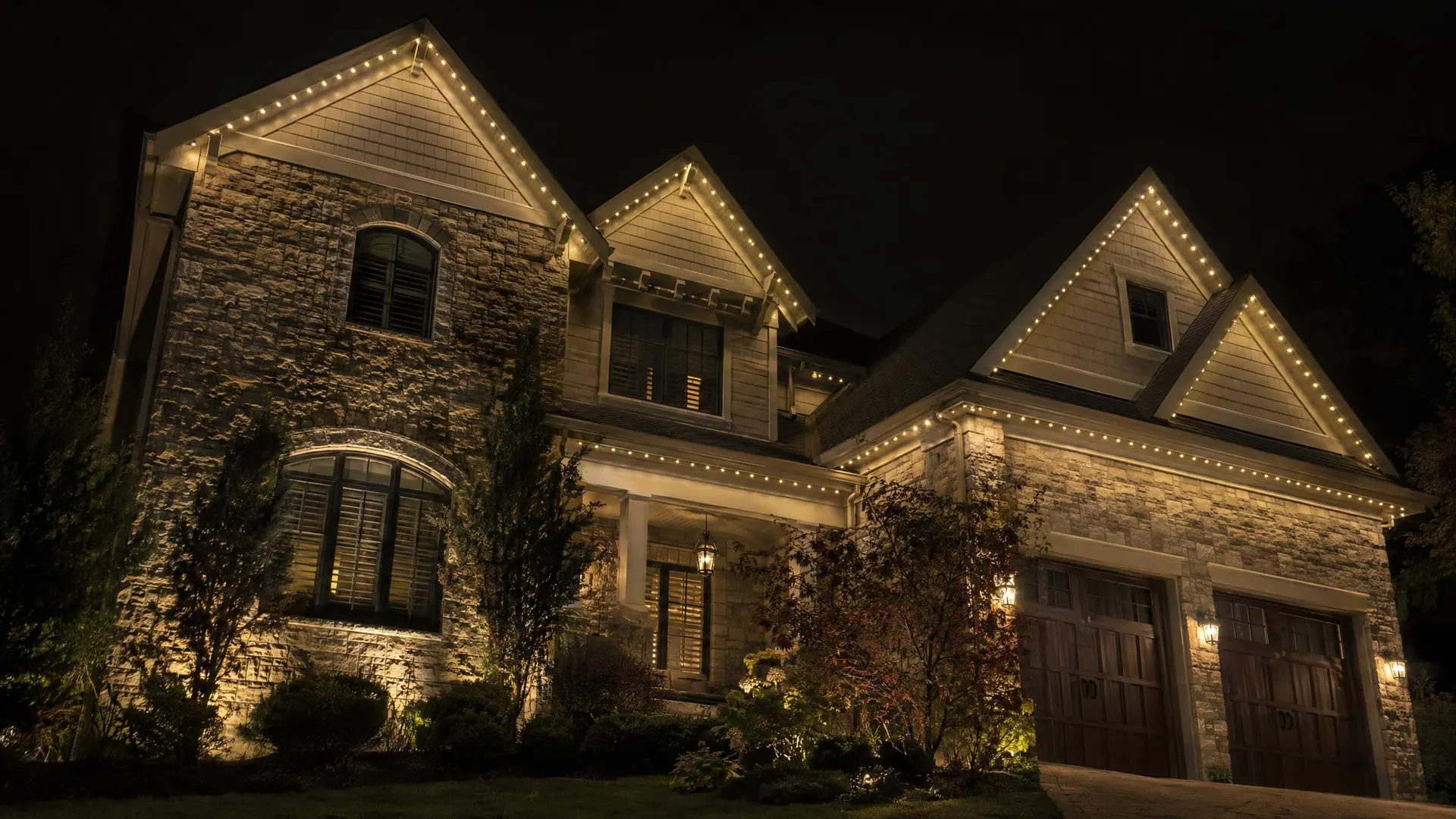 A stone-and-shingle home seen from a low street-level angle at night, roofline in warm-white light