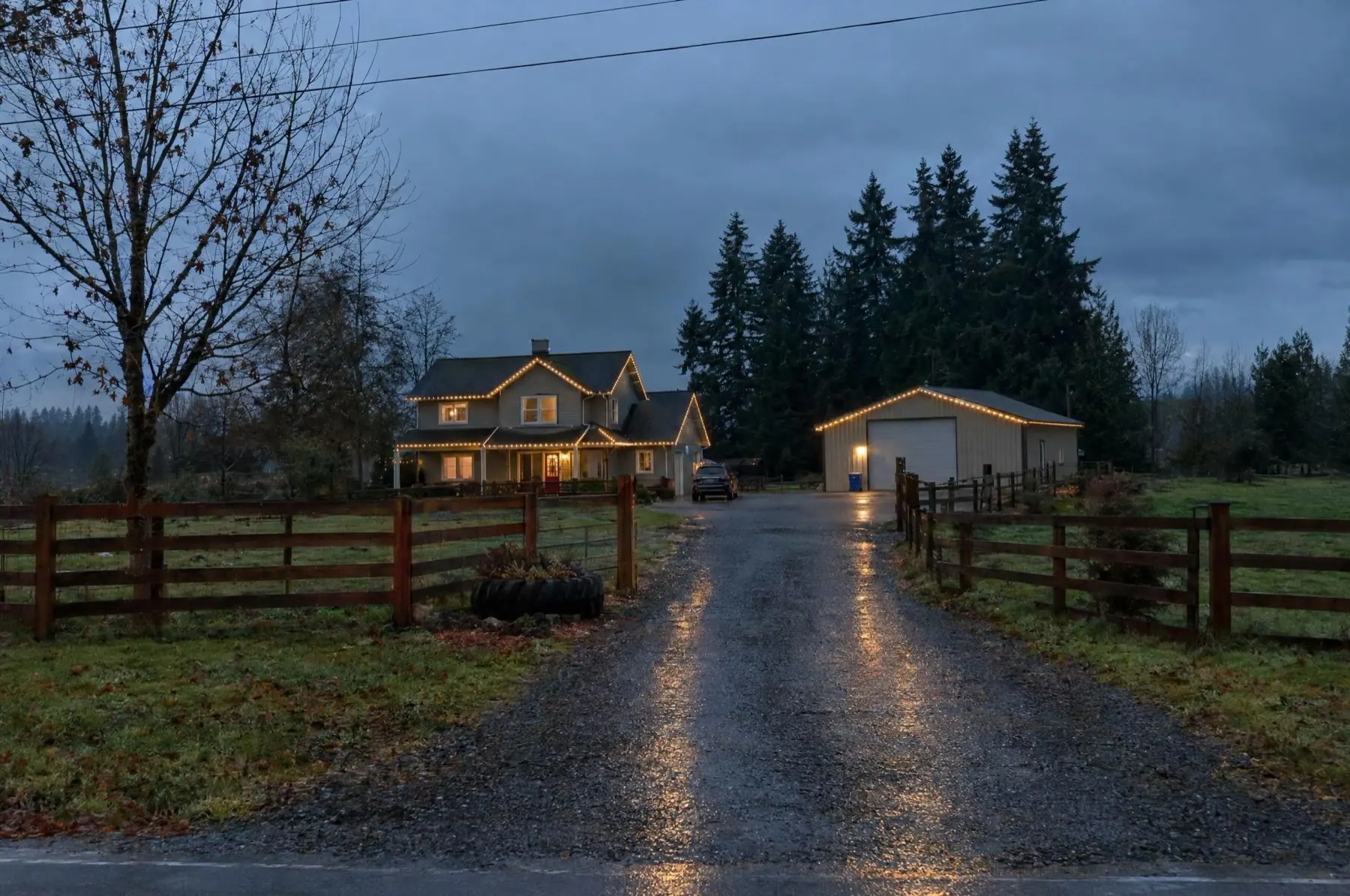 Farmhouse and steel shop both lit warm white at the end of a gravel driveway